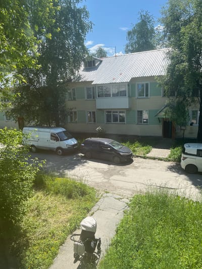 A medium shot captures an overcast, daytime scene in Baykalsk, Russia, focusing on a two-story apartment building with a corrugated metal roof. The building's facade is a combination of light yellow and pale green siding. In front of the building, a grey asphalt road is visible, with parked vehicles including a white van, a dark-colored sedan, and the rear of a white compact SUV. A stroller is positioned on a cracked concrete pathway near a grassy area with yellow wildflowers. Trees with dense green foliage partially obscure the building and the sky. The overall impression is a residential, urban environment on a bright day.