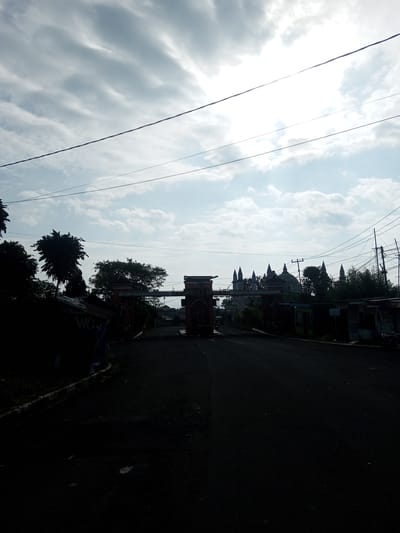 The image depicts a street in Mya Sit Kaing Ywar Thit, Myanmar, leading towards an ornate gate. Beyond the gate, the silhouettes of several structures with spire-like rooftops, possibly part of a religious building or temple, are visible. Power lines crisscross the sky above. The foreground of the street is largely obscured by shadow, while structures line either side, including what appear to be small shops or stalls, one with the letters "WC" visible. The sky is cloudy with a bright area in the middle, possibly the sun filtered through the clouds.