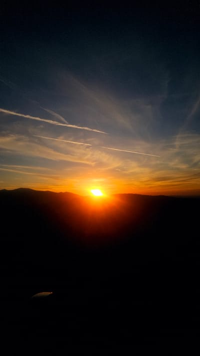 The image depicts a sunset over a mountainous landscape. The sun, a bright, intense yellow-orange orb, is setting behind a dark silhouette of hills, casting a warm glow across the sky. Wispy clouds, tinged with orange and pink, stretch across the horizon, some appearing as contrails from aircraft. The sky transitions from a deep blue at the top to vibrant oranges and yellows near the horizon. The foreground is a dark, silhouetted expanse, likely the side of a hill or mountain, with a faint, reflective object visible on the lower left. The scene is serene and evokes a sense of natural beauty and the end of the day. The location is identified as Simeonovets, Bulgaria.