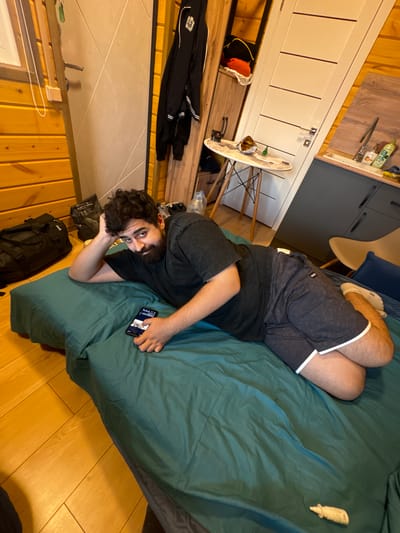 A man with a beard is lying on a bed with teal bedding, resting his head on his left arm. He is wearing a dark t-shirt and patterned shorts. In his right hand, he holds a smartphone, which displays an image. The bed is positioned on a wooden floor. To the left of the bed is a black duffel bag. In the background, there is a wooden wall, a white door, and a small table with items on it. The environment appears to be an indoor residential setting, potentially a room in a house or apartment.