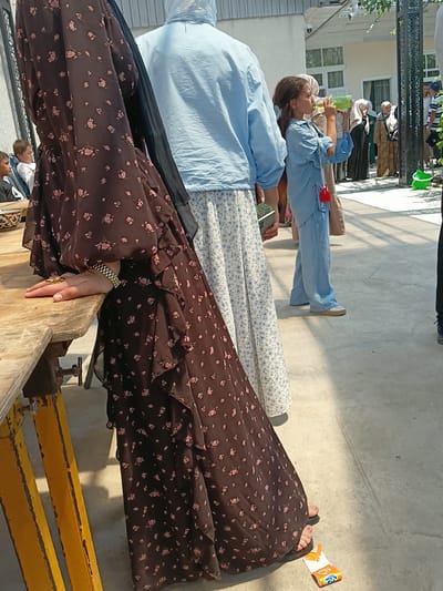 The image depicts a street scene in Osh, Kyrgyzstan, on a sunny day. In the foreground, a person wearing a dark brown, floral-patterned dress with ruffles is leaning on a wooden table with yellow supports. The person's arm, adorned with a bracelet and rings, rests on the table. To the right of this individual, a young girl in a light blue outfit is drinking from a green bottle. Further in the background, several other people are visible, some wearing headscarves and traditional clothing, suggesting a gathering or public event. The environment appears to be an outdoor area with a paved ground surface and some architectural elements in the background, including windows and a metal gate.