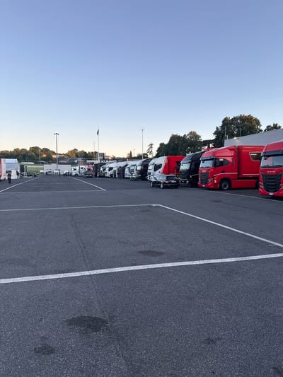 The image shows a parking lot filled with parked trucks in Campagnano di Roma, Italy. The trucks are lined up in rows, mostly red and white. A Kumho tire sign is visible. The parking lot is paved with asphalt and marked with white lines. The sky is clear and blue. Trees line the edge of the parking lot.