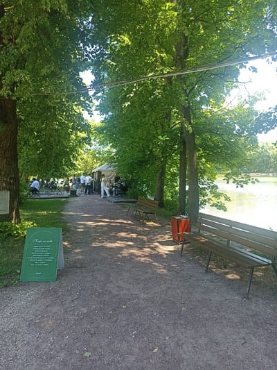 In Ostafievo, Russia, a serene outdoor cafe setting unfolds along the edge of a tranquil body of water. The scene is framed by lush, verdant trees, their leaves dappled with sunlight filtering through the canopy. A gravel path winds through the area, leading to several tables and chairs where patrons are seated, enjoying the ambiance. To the right, a wooden bench offers a place to rest, with a red trash receptacle nearby. Further in the distance, a tent-like structure serves as a backdrop for the cafe's operations, with a few individuals engaged in conversation. The water's surface, reflecting the sky and trees, adds to the peaceful atmosphere of this outdoor gathering space.