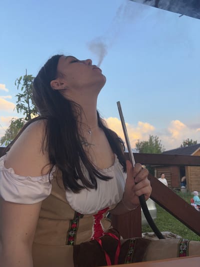 A woman is smoking a hookah outdoors in Пальники, Russia. She is wearing a traditional white and brown embroidered dress. Smoke emanates from her mouth as she holds the metal hookah pipe. The sky is visible in the background with some light clouds. Parts of a wooden structure are also visible. Other people can be seen in the background, possibly sitting at tables.