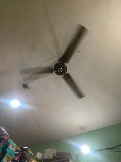 The image shows a ceiling fan in a room located in Kanpur, India, at coordinates 26.44 latitude and 80.344 longitude. The fan is brown with three blades and is spinning. The blades are blurred due to the motion of the fan. The fan is attached to a white ceiling. There are two light fixtures in the ceiling, both of which are on. The room appears to be a shop or a store, with packages of snack food visible in the bottom of the frame. The walls are painted a light green color. The ceiling is a textured white. There is no furniture or people in the room. The fan is the primary subject in the image. The light fixtures, the walls, the ceiling, and the packages of food are all secondary subjects in the image. The image was taken from a low angle. The camera is pointed upward toward the ceiling.