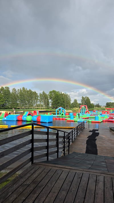 The image shows a vibrant inflatable water park on a lake under a cloudy sky with a prominent double rainbow. In the foreground, a wooden dock with dark railings extends into the water. Scattered across the lake are numerous colorful inflatable structures including slides, climbing walls, and floating platforms. Several people are visible on the inflatables, interacting with the equipment. The background features a line of green trees and vegetation. The scene is located in Jaunmārupe, Latvia, likely at a recreational water park.