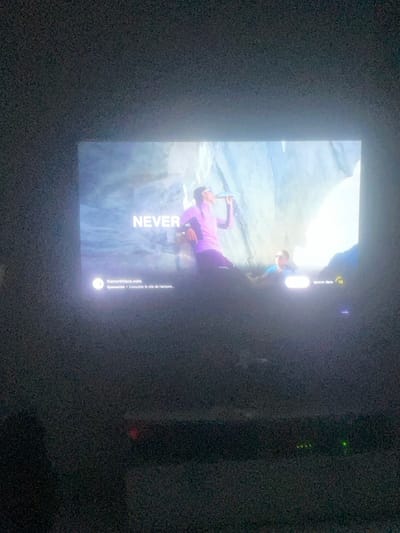 The photograph, captured in a dimly lit room in Vendenheim, France, displays a screen showing an advertisement. The screen content features two individuals within a light-colored, rocky, cave-like environment, illuminated by bright natural light originating from the right side of the frame. The primary subject, positioned left of center, wears a purple long-sleeved top, dark pants, and white-framed sunglasses, and is actively drinking from a cylindrical bottle. A second individual, wearing a blue top and sunglasses, is partially visible in the lower right background, appearing to recline against the rocky terrain. The word "NEVER" is displayed in large white capital letters on the left side of the screen. Below this text, the website "thenorthface.com" is visible, accompanied by a small white circular icon and French text indicating it is a sponsored advertisement. On the lower right, a white box with French text "Ignorer dans" (Ignore in) is present, along with a yellow circular icon containing the number "13," signifying a countdown. Below the screen, in the dark foreground of the room, a horizontal dark electronic device, likely a soundbar or media console, is visible, displaying several small green indicator lights.