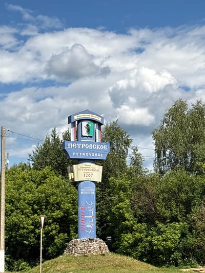 This is a photograph of a village sign in Petrovskoye, Russia. The sign is tall and multi-faceted, with various inscriptions and emblems. At the top, a blue banner with white text reads "Ярославская область," indicating it is located in the Yaroslavl Oblast. Below this, a smaller blue sign features a coat of arms with a black silhouette of a man on a horse against a green and white background. The main body of the sign displays "ПЕТРОВСКОЕ" in large, blue, Cyrillic letters, with "PETROVSKOE" in English below it.

To the right of this central section, a beige sign provides historical information about the village, stating "Первое упоминание о селе ПЕТРОВСКОМ Датируется 1207 годом," which translates to "The first mention of the village PETROVSKOM dates back to 1207." Below this, a blue banner with white text reads "ДОБРО ПОЖАЛОВАТЬ" and "WELCOME!", signifying a friendly greeting to visitors. Further down on the blue sign, there are stylized red lines and the text "M8" and "ЯРОСЛАВЛЬ," suggesting proximity to the M8 highway and the city of Yaroslavl.

The sign is set against a backdrop of lush green trees and a bright blue sky with white, fluffy clouds. The overall scene appears to be a sunny day, likely during daylight hours. The sign itself is constructed with a mix of materials, including what appears to be stone at the base. There are no people visible in the photograph. The image is oriented vertically.