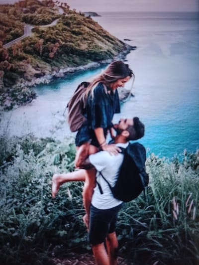 A man is holding a woman in his arms while standing on a grassy hill overlooking a body of water. The man is wearing a white t-shirt and dark shorts, and a black backpack. The woman is wearing a dark top and a light-colored backpack, and has her legs wrapped around the man. The background shows a coastline with lush green vegetation and a winding road. The water is a vibrant turquoise color, and the sky is a soft purple and blue gradient. The overall scene depicts a romantic and adventurous moment between a couple in a scenic natural setting. The location is not Balakliya, Ukraine; it is a coastal mountainous area with abundant greenery and ocean views.