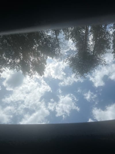 The image shows a view upwards through what appears to be a sunroof or open window, looking at a sky with trees partially obscuring the view. The sky is blue with scattered white clouds. The tops of trees, silhouetted against the sky, are visible in the upper portion of the image. The scene is likely outdoors and the lighting suggests it is daytime. There is no discernible location in the city of Molodyozhnoye, Russia, visible or implied in this image.
