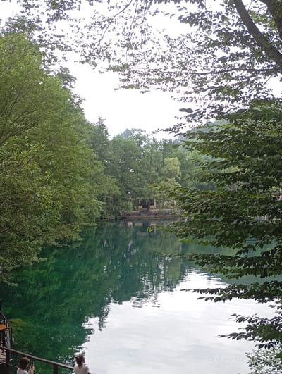 The image depicts a serene natural scene with a clear, turquoise body of water reflecting the lush green foliage of surrounding trees. The water's surface is calm, with gentle ripples distorting the reflections of the trees and a few buildings visible in the distance. The trees form a dense canopy, with some branches extending into the foreground, framing the view of the water. The sky is overcast, casting a soft, diffused light over the scene. In the lower left corner, two individuals are visible, standing on what appears to be a wooden deck or walkway, with one person holding a phone. The timestamp "26.07.2026 15:57" is superimposed on the lower right corner of the image. The scene is consistent with the natural beauty of Verkhnyaya Balkariya, Russia, known for its picturesque landscapes and clear mountain lakes.