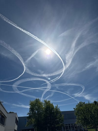 The image displays a bright, sunny sky with numerous contrails from airplanes forming various circular and crisscrossing patterns. The sun is visible, creating a lens flare effect in the upper right quadrant. Below the sky, the upper portions of several modern buildings are visible, along with lush green trees. The architectural style of the buildings suggests an urban or suburban setting, likely in a developed area. The presence of trees and modern structures indicates a location with both natural elements and human infrastructure. The overall scene is one of a clear day with evidence of air traffic. This photograph was taken in Salach, Germany.