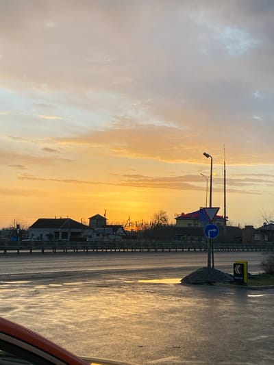 An eye-level shot captures a roadside view at sunset. The sky transitions from pale blue-grey to a radiant golden hue. Road signs and streetlights stand beside a wet road reflecting the sky. Buildings are silhouetted against the bright sky. A sliver of a vehicle's exterior appears at the frame's edge.