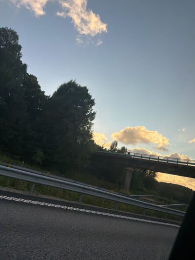The image depicts a view from a vehicle traveling on a highway near Ödsmål, Sweden. The scene is characterized by a clear blue sky with scattered, brightly lit clouds, suggesting it is either early morning or late afternoon. On the left side of the frame, dense, dark green trees line a hillside, with their tops partially obscuring the sky. In the middle ground, a concrete overpass with a light blue railing is visible, supported by a single concrete pillar. The overpass stretches across the frame, and beyond it, the terrain is covered in trees and vegetation. The highway in the foreground has dark asphalt and is marked with double white lines. A metal guardrail runs along the edge of the highway. The overall impression is of a scenic drive through a forested area under a dynamic sky. There are no people or animals visible in the image.