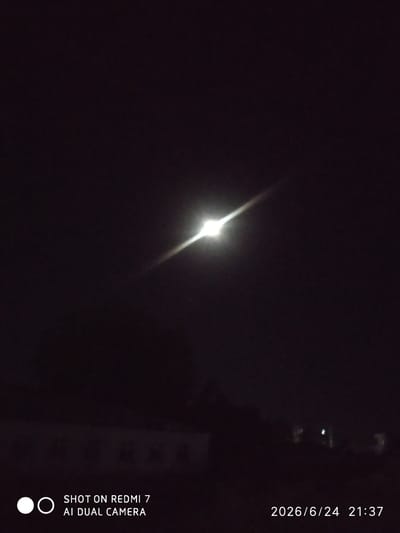 This image depicts a nighttime scene in Khalkabad, Uzbekistan. The primary subject is a bright, artificial light source, likely a streetlamp, positioned in the upper center of the frame. The light is intensely luminous, creating a starburst effect and casting a diagonal ray of light across the dark sky.
In the lower portion of the image, a faint, blurry outline of buildings or structures is visible against the dark background. The overall setting is that of a quiet, possibly rural or suburban area at night, with minimal ambient light apart from the dominant lamp.
The time indicated by the visible text is 21:37 on June 24, 2026. The sky is completely dark, suggesting a clear night with no significant cloud cover. There are no visible people or animals. The mood is subdued and solitary, dominated by the stark contrast between the artificial light and the surrounding darkness.
Visible text on the image includes:
- "SHOT ON REDMI 7"
- "AI DUAL CAMERA"
- "2026/6/24 21:37"