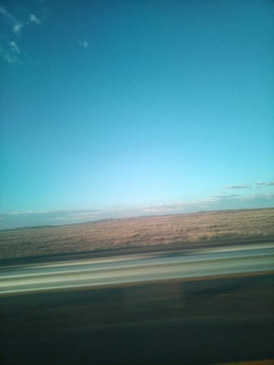 This media file captures a vast, open landscape under a clear, light blue sky, indicative of daytime in a dry, possibly arid region. The scene appears to be viewed from a moving vehicle, evidenced by the motion blur in the foreground and along the horizontal lines of what looks like a road or highway.
The primary subject is the expansive, flat terrain stretching towards a low, distant horizon. The ground is covered in dry, muted-toned vegetation, possibly grass or scrub, with subtle variations in color from tan to light brown, suggesting a natural, undeveloped setting. Faint, undulating hills or mountains are barely discernible in the far distance, blending into the horizon.
Several horizontal streaks of light and color dominate the foreground. These appear to be the result of motion blur, possibly representing road markings or reflections from the vehicle's interior or the road itself. The colors are predominantly light green, white, and yellow, with darker tones below.
The sky is a dominant feature, occupying the upper two-thirds of the frame. It is a clear, bright blue, with a few wispy, white clouds scattered sparsely near the horizon, particularly on the left side. The overall lighting suggests it is a bright day, likely late morning or early afternoon, given the clarity of the sky and the absence of strong shadows.
The location context provided is Aktau, Kazakhstan, which aligns with the visual cues of a vast, sparsely vegetated, and potentially desert-like landscape. There are no people or man-made structures clearly visible, emphasizing the sense of open space and natural environment. The emotion conveyed is one of vastness, movement, and perhaps a sense of journey or travel through a seemingly endless landscape. No text is visible in the media file.