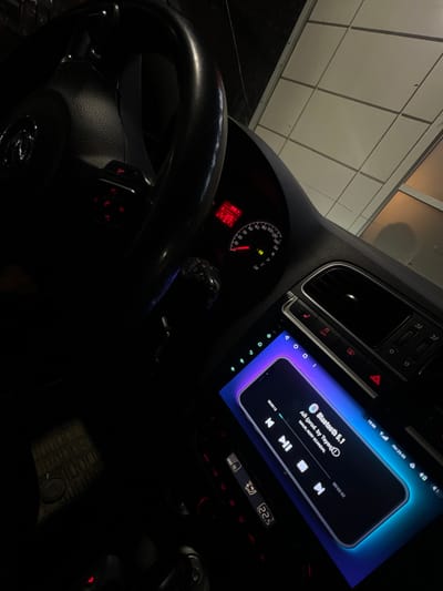 The image captures the interior of a car, likely taken from the driver's perspective looking towards the right side of the dashboard and center console. The scene is dimly lit, suggesting it's either nighttime or the car is in a parking garage, with overhead artificial lighting reflected on the windshield or dashboard.

In the foreground, the dark steering wheel is prominently featured, with red illuminated controls visible on its right spoke. A Volkswagen (VW) logo is partially discernible at the bottom center of the steering wheel. To the left, behind the steering wheel, part of the instrument cluster is visible, showing a speedometer dial with markings up to 240 km/h, and a needle indicating a speed between 0 and 40 km/h (likely around 20-30 km/h). A digital display within the instrument cluster shows red text, possibly indicating time, temperature, or a warning (a red icon resembling a battery or engine light is also present).

The central focus of the image is a large, rectangular infotainment screen integrated into the car's center console. This screen displays a music player interface, resembling a smartphone's media playback screen. The top left of the player shows "Bluetooth 5.1," indicating the connection type. The currently playing track is "AR (prod. by Teyeq)," with artists listed as "MAUR, KERY SCANDAL." A progress bar shows "00:00:12" as the current timestamp and "00:02:52" as the total duration of the song. Playback controls (skip backward, play/pause, skip forward) are clearly visible at the bottom of the player interface. The top bar of the infotainment screen displays the time "19:58" and the date "ПН 23.02" (Monday, February 23rd, with "ПН" being the Cyrillic abbreviation for Monday). Other icons on this bar suggest signal strength, Wi-Fi, Bluetooth, and battery status.

Below the infotainment screen, the car's climate control panel is visible, displaying "22.5," likely indicating the temperature setting in degrees Celsius. Buttons for hazard lights (red triangle), central locking, and an "OFF" button (possibly for traction control or another system) are also visible on the center console. Air vents are present above these controls. The overall impression is that music is being played inside a parked or slowly moving car in a low-light setting.