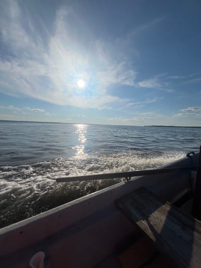 The image captures a view from inside a small boat moving across a large body of water, likely a lake or a calm sea. The sun is high in the sky, bright and positioned to the left, casting a strong glare on the water's surface and creating a shimmering reflection. Scattered white clouds dot the bright blue sky. The water is rippled, with a visible wake trailing behind the boat, indicating forward motion. A dark-colored oar or tiller extends across the foreground, partially submerged in the wake. The interior of the boat is visible in the lower right portion of the image, revealing a dark hull and a wooden plank. In the distance, a faint, tree-lined horizon is visible. The scene suggests a sunny day, possibly in the summer, experienced from a boat on a body of water near Siukterka, Russia.