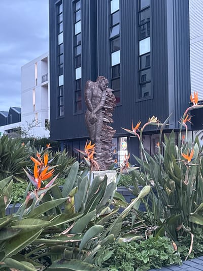 A street-level, slightly blurred photo shows a cityscape with tropical foliage in the foreground. Orange Bird of Paradise flowers are prominent. Behind the flowers, there's a building with horizontal, windowed layers. A stone sculpture or abstract statue is positioned in front of the building. A white building is visible lower down. The image appears to be taken from ground level, focusing on the greenery and structures along a street in Milnerton, South Africa.