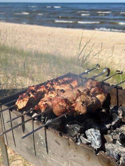 A close-up shot captures skewers of meat grilling over hot coals. The meat is well-seared, with some pieces showing char marks from the intense heat. The skewers are arranged in a metal grill that sits on a concrete surface. In the background, out of focus, is a sandy beach with some dry grass. To the left, a body of water, likely the sea, is visible, with gentle waves lapping at the shore. The lighting suggests it is daytime, possibly late morning or early afternoon, with the sun casting shadows on the grill and the ground. The overall impression is one of a relaxed outdoor barbecue, perhaps at a seaside location. There is no visible text in the image.