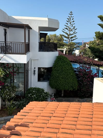 The image displays a bright, sunny day with a clear blue sky above. In the foreground, a tiled terracotta roof slopes downwards. Behind the roof, a modern white building with balconies and dark railings is visible. Lush green plants and flowering bushes, some with pink and white blooms, surround the building. A swimming pool with blue water is situated in the mid-ground, partially shaded by an awning. In the background, more trees and vegetation are seen, with a glimpse of the sea and a distant coastline. The scene appears to be a resort or hotel in Chersonisos Municipal Unit, Greece, characterized by Mediterranean architecture and landscaping.