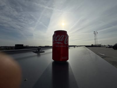 A red Coca-Cola "Original Taste" aluminum can stands upright on a wide, flat, light-grey surface, likely the rooftop of a parking garage, at the Hoogvliet De Lier retail complex, located at Van Rijckevorselstraat 12, 2678 CK De Lier, Netherlands. The can is positioned slightly left of center, casting a distinct shadow to its right due to strong backlighting from the sun, which is visible as a bright orb in the upper-center sky. The sky is partly cloudy with multiple linear contrails.
To the left of the can, on the same flat surface, is a small, metallic square base plate with a cylindrical protrusion. In the lower-left corner, a blurred, tan-colored human digit partially obscures the view.
Beyond the foreground surface, the scene extends to an urban landscape. To the left, distant low-rise industrial or commercial buildings with dark facades are visible under a hazy horizon. To the right, the flat surface transitions into an asphalt parking lot marked with white directional arrows, where several dark-colored vehicles are visible in the distance. A tall, slender, lattice telecommunications tower, equipped with multiple antenna platforms, stands prominently in the mid-ground against the sky, to the right of the Coca-Cola can. No individuals are directly visible in the scene, and no specific actions or interactions are depicted beyond the static placement of the can.