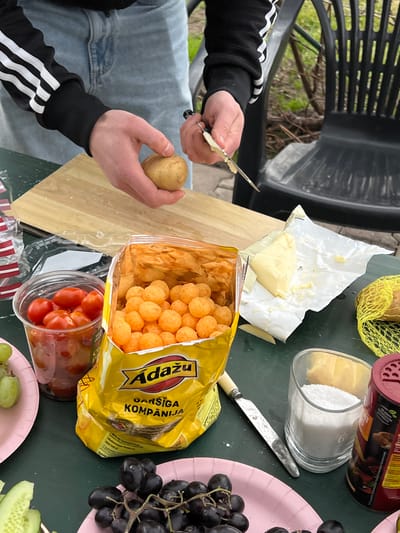 A person with a black and white Adidas tracksuit is slicing a potato on a wooden cutting board. On a green surface is a yellow bag of "Adažu" brand cheese balls, along with butter, a glass jar filled with white powder, a jar of chili powder, and several small containers of vegetables. A pink paper plate of grapes and cucumbers is on the right of the frame. The image suggests a picnic or outdoor food preparation setting. Dārzciems, Latvia is not confirmed.