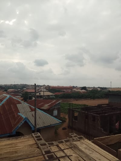 An elevated, wide-angle photograph captures a developing urban landscape in Arun, Nigeria, under a cloudy, grey sky. The immediate foreground shows an active construction site featuring wooden planks, hollow concrete blocks, and exposed rebar, indicating the image was captured from an unfinished upper level. Below and to the left, two completed residential buildings are visible; the closer one has a rusty, reddish-brown corrugated metal roof with light blue eave trim, while the one behind it also features a rusty metal roof. A weathered, dark wooden pole ascends vertically from this area. The midground consists of a large section of reddish-brown disturbed earth, bordering a construction zone on the right that includes the skeletal frames of two incomplete concrete block structures with exposed rebar. A partial reddish sign is visible on the far-right incomplete structure. Green vegetation, including bushes and trees, delineates the boundary between the undeveloped land and the background. The background reveals a dense residential district extending to the horizon, comprising numerous buildings with varied roof types. A tall telecommunication mast is present on the far-right horizon. No individuals are discernible within the frame.