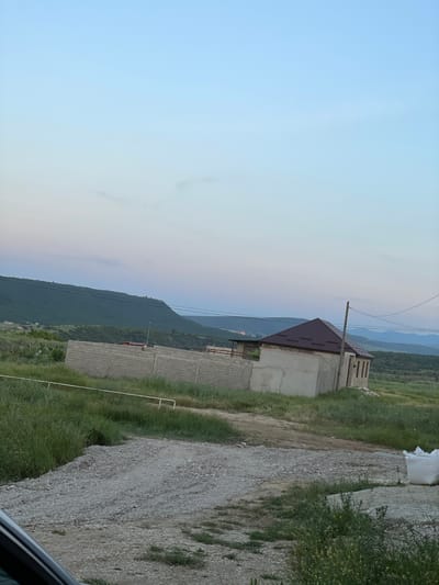 The image depicts a rural landscape near Buynaksk, Russia, under a pale, twilight sky. In the foreground, a dirt and gravel road leads towards a small, unfinished building with a dark, sloped roof. The building is constructed of concrete blocks and has several visible window openings. Adjacent to the building is a low wall made of grey concrete blocks, extending horizontally across the midground. Beyond the wall, rolling green hills and mountains form the backdrop, with the larger mountains in the distance exhibiting a hazy, bluish hue. The ground is covered in grass and some sparse vegetation. A white bag, possibly containing construction materials, is visible to the right of the road. The scene is devoid of any people or animals.
