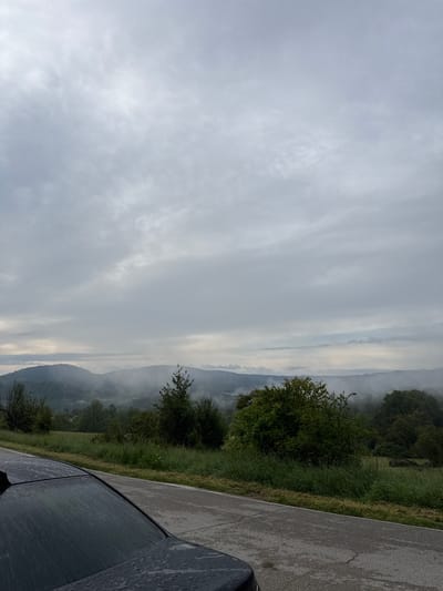 A misty, overcast day is depicted in this image, with a wide view of rolling hills shrouded in fog. The foreground shows the wet roof and side of a dark-colored vehicle, suggesting recent rain. A paved road runs alongside a grassy embankment, leading towards the hazy landscape. The sky is filled with heavy, gray clouds, contributing to the muted and serene atmosphere of the scene. The location is identified as the city of Boltata, Bulgaria. There are no people or animals visible in the image.