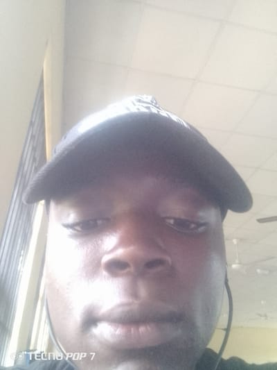 A close-up, downward-angle selfie shows a dark-skinned individual wearing a dark baseball cap. The person's face is illuminated by bright overhead lighting, creating a shiny effect on their skin. Their eyes are partially closed, and their mouth is slightly open, revealing teeth. Visible on the left side of the image is a portion of a metal grate or railing, suggesting an indoor setting with some natural light filtering in. In the background, a white ceiling with visible tiles and two ceiling fans are present. The bottom left corner of the image displays the watermark "TECNO POP 7". The overall impression is that of a casual, indoor photograph, possibly taken in a public space or common area.