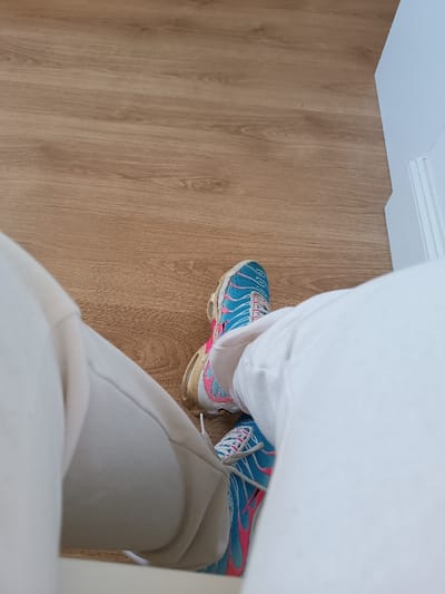 The image presents a first-person perspective looking down at a person's legs and feet, featuring a pair of distinct sneakers. The person is wearing light-colored, possibly off-white or cream, comfortable trousers or sweatpants.
The central subjects are a pair of athletic shoes, specifically Nike Air Max Plus (TNs), which are brightly colored in shades of blue and pink. The main body of the shoes is turquoise blue, adorned with prominent pink, flame-like wavy patterns on the sides. The laces are white, and the soles are a yellowish, somewhat aged or discolored translucent material, suggesting wear. A white Nike swoosh logo is visible on the side of one shoe.
The setting appears to be indoors, likely a room with a wooden floor. The floor consists of light brown, horizontally laid planks, showing a visible wood grain texture. To the right side of the frame, a segment of a white wall or baseboard is visible, suggesting a clean, minimalistic interior space.
No specific activity beyond the person being seated or having their legs extended is discernible. There are no visible details indicating the time of day, weather, or emotions. No explicit text other than the Nike logo is clearly legible. The broader context for the media file is Thionville, France, though there are no visual cues within the image to specifically identify this location.