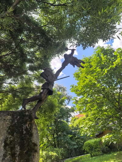 The image shows a bronze statue of a man in historical attire, possibly a hunter or warrior, in mid-leap. He is reaching up to grasp another figure, which is a child, also in mid-air. The statue is positioned on a rough, moss-covered stone pedestal. Lush green trees with dappled sunlight filter through the leaves, creating a dynamic interplay of light and shadow. In the background, a bright blue sky with scattered white clouds is visible, along with hints of buildings and more foliage, suggesting an outdoor setting. This statue is located in the park of the Sovkhoza imeni Lenina, Russia.