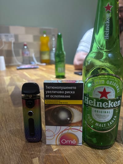 A still life photograph is presented, featuring a Heineken beer bottle prominently on the right side of the frame. Next to it, to the left, is a pack of cigarettes, and to the left of the cigarettes is a vape device. The items are arranged on a wooden surface. In the background, blurred out, are various bottles and what appears to be a kitchen counter with electrical outlets. The lighting suggests an indoor setting, possibly a kitchen or dining area. There is no indication of the city of Boston, United Kingdom.