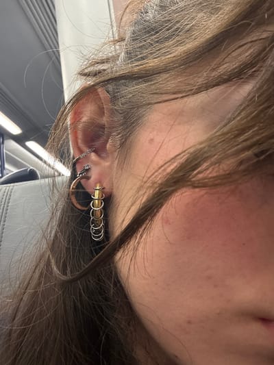 A close-up, sideways view of a person's ear reveals multiple piercings adorned with various jewelry. An upper cartilage piercing features a bar with small, shiny gems. A lower piercing displays a crescent-shaped hoop. The lobe showcases a series of rings, including a golden one followed by several silver ones linked together, creating a chain-like effect. The person has light skin and brown hair. The background includes a glimpse of what appears to be a train seat and window.