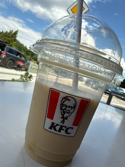 A close-up, outdoor shot focuses on a KFC branded beverage cup filled with a light-colored, creamy liquid and topped with a clear plastic dome lid and a gray straw. The KFC logo, featuring Colonel Sanders, is prominently displayed on the cup. The cup rests on a white surface, possibly a table. In the blurred background, a dark-colored SUV is visible with its rear door open, parked on a paved area adjacent to a road. Further in the distance, a yellow diamond-shaped road sign is partially visible against a blue sky with scattered clouds. The overall scene suggests an outdoor setting, possibly a roadside cafe or rest stop in the vicinity of Kalugerovo, Bulgaria. There are no discernible people or direct interactions captured in the frame.