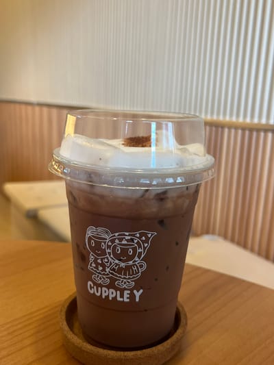 A chocolate beverage in a plastic cup with a clear lid sits on a light brown, cork-like coaster. The cup is dark brown with a cartoon graphic and the word "Cuppley" printed in white. The drink has a layer of white topping, possibly whipped cream, visible through the lid. The background features light brown horizontal stripes and a light beige surface, likely a table. The image was taken in the city of Muang Pattaya, Thailand.