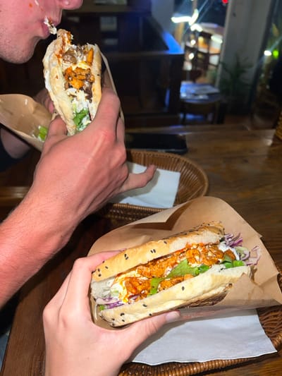 The image depicts an indoor, nighttime scene within an eatery in Thành phố Phan Thiết, Vietnam, where two individuals are consuming banh mi sandwiches. In the upper left, a person is shown actively biting into a banh mi, with white sauce residue visible on their chin. Their left hand, exhibiting a tanned forearm, grips the sandwich, which is partially wrapped in brown paper and reveals a filling of meat or tofu, lettuce, and sauces. In the lower right, a second individual holds a complete banh mi in their left hand. This sandwich features a baguette-style roll topped with black sesame seeds, filled with orange-toned protein, green lettuce, shredded red cabbage, and white and orange sauces, also enclosed in brown paper. Both sandwiches are presented in separate brown rattan baskets, each lined with a white paper napkin. A dark-colored smartphone rests on the wooden table surface between the individuals. The background is blurred, displaying warm ambient lighting and indistinct wooden furniture, indicative of a casual dining establishment.
