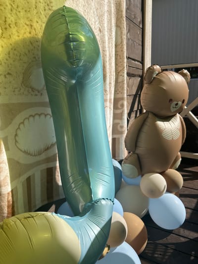 An outdoor scene features a collection of inflated balloons arranged as a decoration. Dominating the foreground is a large, turquoise, vertically oriented balloon that appears to be an elongated number or shape, casting a shadow on a patterned fabric backdrop. To the right of the turquoise balloon is a brown, inflatable teddy bear with subtle facial features and a checkered bowtie. The bear is seated on a cluster of smaller, round balloons in various pastel shades of blue, cream, and brown. The balloons rest on a wooden deck, with a blurred wooden structure and beige curtains visible in the background. The lighting suggests it is a sunny day, with dappled sunlight and shadows. The location is specified as the city of Lyamino, Russia. There are no people present in the image, nor are any interactions depicted.