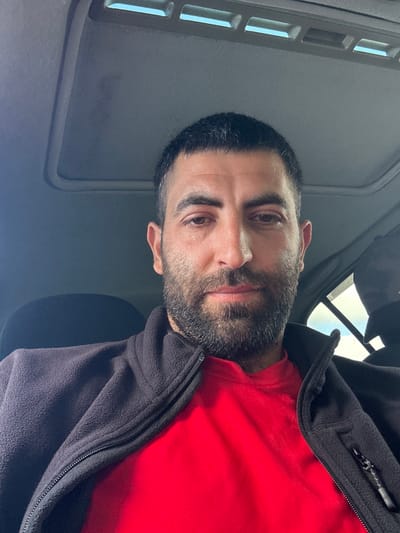 A man with short dark hair and a full beard is seated inside a vehicle, taking a selfie. He is wearing a red t-shirt and a dark grey fleece jacket. The interior of the car is visible, including the roof lining with a sunroof. The background suggests an outdoor environment with daylight. There is no indication of the location being in Adur, United Kingdom.