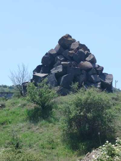A towering, conical pile of dark, irregularly shaped rocks dominates the center of the frame, situated on a grassy hillside under a clear blue sky. The rocks are of various sizes, with some appearing rough and others smoother. The hillside is covered in green grass and scattered with small, leafy bushes. To the left of the rock pile, a thin, bare-branched tree stands out against the sky. In the distance, a grey structure, possibly a bridge or elevated roadway, is visible on the left side of the image. On the far right, a thin pole with what appears to be a light fixture or sensor is present. The foreground shows more dense green vegetation and a patch of white flowers on the right. The scene appears to be located in a natural, outdoor environment, likely in the vicinity of Jrvej, Armenia, given the presence of basalt rock formations characteristic of the region. There are no people or animals visible in the image.