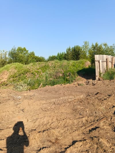 The image captures a bright, clear day with a prominent blue sky dominating the upper portion. In the middle ground, a grassy embankment rises, topped with a line of green trees and shrubs. To the right of the embankment, a weathered wooden fence, consisting of vertical planks, stands prominently. The foreground is composed of disturbed, brown soil, with tire tracks visible, suggesting recent activity, possibly from construction or landscaping. A shadow of a person holding a camera or phone is cast on the soil in the lower left corner, indicating the photographer's presence. The scene appears to be an outdoor, possibly rural or semi-rural area, likely on the outskirts of Strelka, Russia, given the presence of natural vegetation and the rough terrain.
