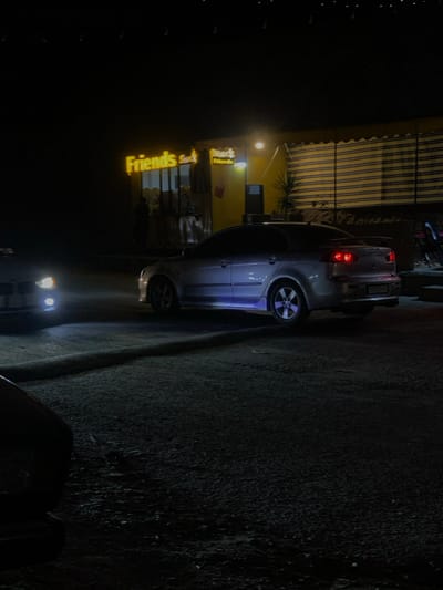 A silver car is parked at night near a building with an awning. The building has a sign that reads "Friends Cafe". Headlights from another vehicle illuminate the street in the foreground. The road surface is dark and textured. Overall, the image is dark with artificial lighting.