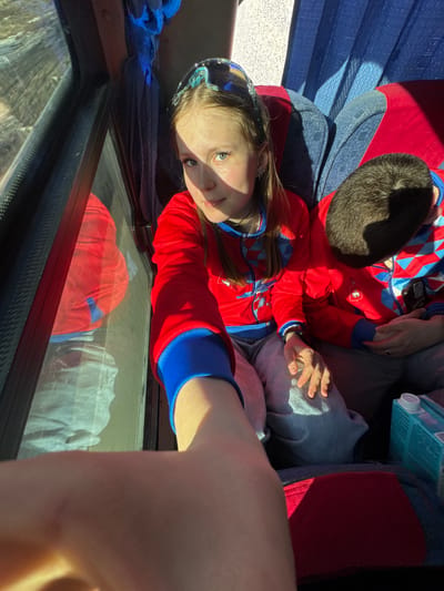 A young girl with light blonde hair and blue eyes is looking at the camera. She wears a red jacket with blue accents and a patterned headband. She is sitting on a bus. Her legs are stretched out. Next to her is a child with dark hair who is also wearing a red jacket with blue accents. An adult's hand is resting on the child. A carton of juice is near their legs. Through the bus window, the distorted reflection of clothing can be seen. The location is likely in Челва, Russia, due to the apparel.