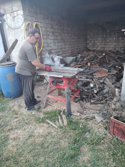 A man wearing glasses, a grey t-shirt, dark pants, and red gloves is standing in front of a red table saw. He is holding a piece of wood and appears to be working with the saw. The saw is set up outdoors in what looks like a backyard or workshop area. Behind the man and the saw is a large pile of logs and wood debris, as well as a blue plastic barrel. The background shows a brick wall and some industrial piping. The overall scene suggests a DIY or woodworking activity taking place in an outdoor, somewhat cluttered environment. This location is identified as being in the city of Kochmar, Bulgaria.