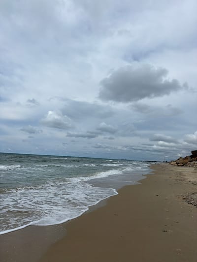 The image displays a coastal scene at Beit Yanai, Israel, under an overcast sky. The upper portion of the frame is dominated by a dense, layered formation of grey and white clouds. The middle ground features the sea, characterized by dark greenish-blue water with multiple white-capped waves breaking towards the shore.
The lower portion of the image shows a sandy beach, light brown to tan in color, extending from the foreground into the distant horizon. A distinct line of white foam marks the immediate interface where waves recede from the wet sand. Numerous footprints are visible in the damp sand along the waterline, indicating human passage. To the right, a low, eroded cliff face composed of light brown, rocky material extends along the coastline into the distance.
On the distant horizon, industrial structures are visible along the coastline. These include a long pier or jetty extending into the sea, with what appear to be ships or large vessels in its vicinity. Further along the coast, several tall industrial chimneys or smokestacks are present, one actively emitting a plume of white smoke or steam that rises vertically before dispersing horizontally. A few small, indistinct human figures are discernible on the distant beach near these structures.
