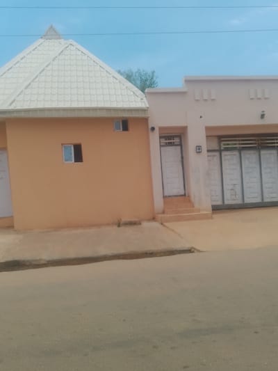 A light peach-colored building with a white, gabled, tiled roof stands to the left of a light tan building with a flat roof. The peach building has a single small, square window with a white frame and blue-tinted glass. The tan building has a white door with a glass panel and metal grate, accessed by a set of three short, wide steps. To the right of the door are two large, multi-panel sliding doors with white frames and glass panels, partially covered by horizontal metal bars. Above these doors, the tan building's facade features three evenly spaced rectangular indentations, two on the left and one on the right. The area in front of the buildings is a sandy, light brown color, sloping down to a paved road. The sky is a pale blue with thin, dark horizontal lines, likely power lines, across the upper portion. The scene appears to be located in Gindi Nassarawa Kyarmi Galbi, Nigeria.