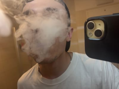 A young adult, with short dark hair and light skin, is exhaling a plume of white vapor. The individual is wearing a white t-shirt and a dark baseball cap. A smartphone with a black case and a triple-lens camera is held up, partially obscuring the right side of the frame. The background appears to be a tiled wall, suggesting an indoor location, possibly a bathroom or shower. The lighting is warm and diffused. The context provided about Gebersdorf, Austria, does not appear to be reflected in the visual elements of the image.