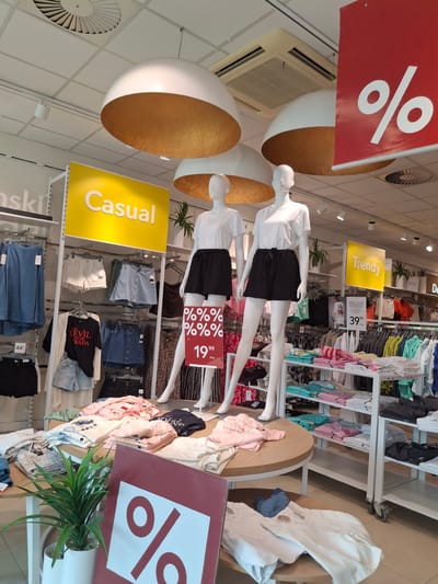 The image depicts the interior of a clothing store in Ząbki, Poland. The store features a bright, modern design with white ceilings, track lighting, and large, dome-shaped pendant lamps with gold interiors. Several mannequins are on display, dressed in casual women's clothing, including t-shirts and shorts. Red signage with large white percentage symbols indicates a sale. Yellow signs on display racks label sections as "Casual" and "Trendy." Shelves are filled with folded clothing items such as t-shirts and shorts, primarily in pastel colors. A potted green plant is placed on a round display table. The flooring appears to be light-colored tile.