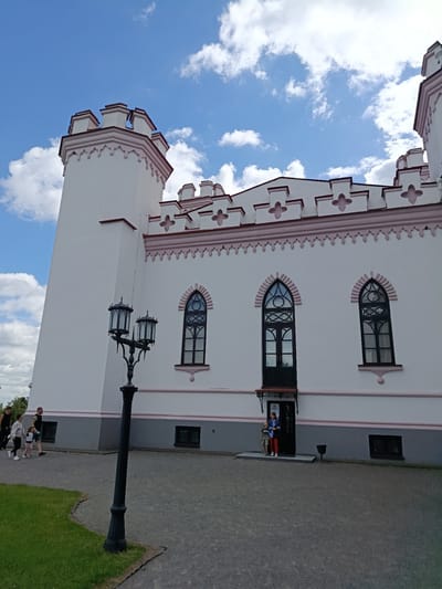 This is a photograph of the Kosava Palace in Kosava, Belarus, under a partly cloudy blue sky. The palace is a white structure with pink decorative trim along its crenellated roofline and around its arched windows. A prominent tower is visible on the left side of the building. In front of the palace, a black, ornate lamppost stands tall, its three lanterns facing outward. Several people are visible on the grounds. On the left, a group of four individuals, including two adults and two children, walk past the building. Near the entrance to the palace, a woman and a young boy stand on the steps. The grounds are paved with gravel, with a patch of green grass to the left.